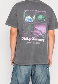 Grå bomull t-shirt med en grafisk ryggdesign som har kosmiska bilder och text i grönt och lila; avslappnad passform med korta ärmar.