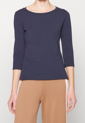 Personne portant un haut bleu marine à manches trois-quarts et un pantalon marron clair avec des bandes blanches sur les côtés sur un fond uni.