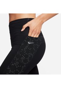 Svarta leggings med hög midja, med en sidoficka och ett grått prickmönster. Nike-logotyp på vänster sida av fickan.