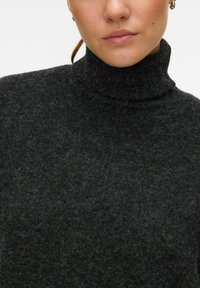 Černý pletený svetr s rolákem a jemně texturovaným povrchem. Má vysoký límec a nenápadné vzory. Detailní pohled na výstřih.
