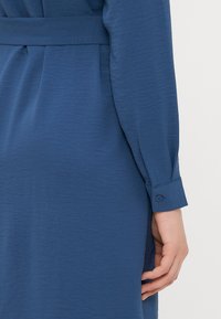 Μπλε φόρεμα με υφή υφάσματος, μακριά μανίκια με κουμπωτές μανσέτες και ζώνη στη μέση για σχήμα.