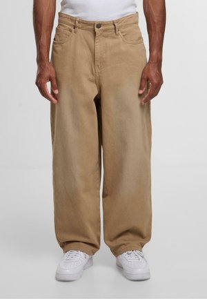 Beige viddebenede bukser lavet af bomuld, med et klassisk fem-lommers design og afsluttet med en knaplukning. Kombineret med hvide sneakers.