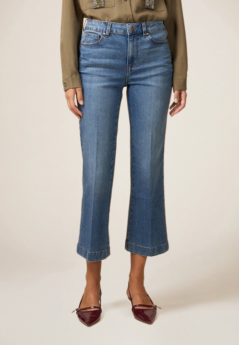 Jeans de denim azul con un diseño acampanado y corto, cintura alta y dobladillo sin terminar. Combinados con una camisa caqui y zapatos burdeos de punta.