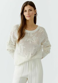 Cremiger Strickpullover mit offener Maschenstruktur, der ein Blattmuster und gerippte Bündchen aufweist. Kombiniert mit hellcremigen weiten Hosen.