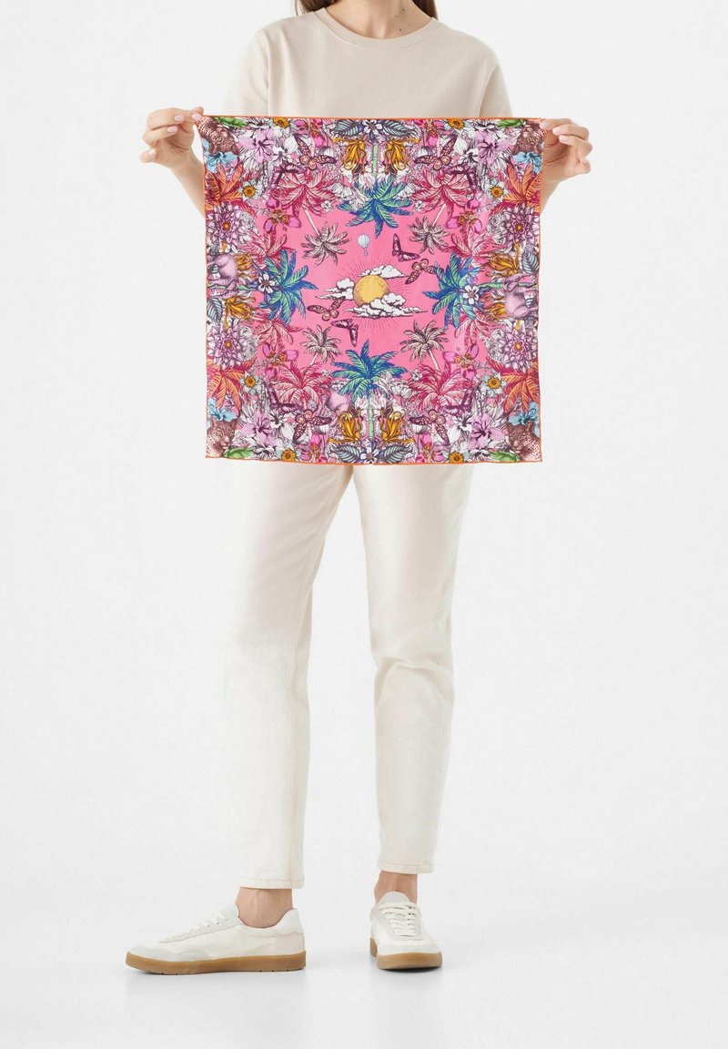 Personne tenant un foulard carré rose avec des motifs tropicaux floraux, palmiers, papillons et soleil complexes.