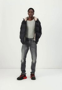 Hombre con chaqueta de plumas negra sobre un suéter gris claro, jeans desgastados negros y zapatillas negras y rojas, de pie contra un fondo blanco.