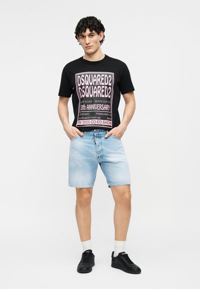 Jeune homme portant un t-shirt graphique noir DSQUARED2, un short en jean bleu clair, des chaussettes blanches et des baskets noires, debout les mains dans les poches.