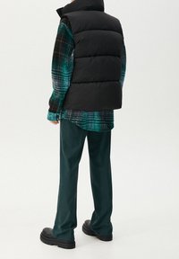 Černá péřová vesta přes modrozelenou kostkovanou košili na knoflíky, doplněná tmavě zelenými kalhotami a černou robustní obuví.