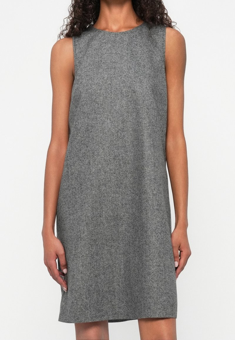 Femme portant une robe en laine grise sans manches, longueur genou, avec un col rond, debout devant un fond blanc.