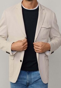 Beiger Blazer aus Stoff mit Revers, einer Brusttasche und Knöpfen an den Ärmelbündchen, über einem schwarzen Hemd getragen. Jeans sind am Boden sichtbar.