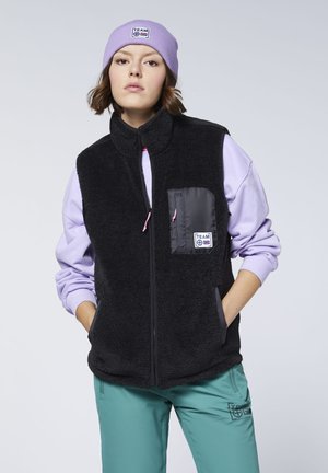 Young woman wearing a black fleece vest, lavender sweatshirt, teal pants, and lavender beanie with "TEAM" logo, standing with hands in pockets.