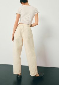 Beige hoge taille wijde jeans met achterzakken, gecombineerd met een lichtgekleurde strakke top en donkere schoenen, tegen een neutrale achtergrond.