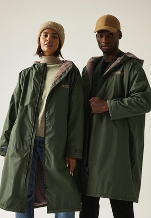Two models wearing oversized olive green lined coats with hoods, one paired with a beige knit hat and the other with a tan cap.