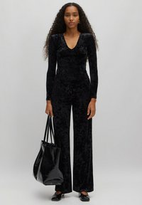 Une combinaison en velours noir avec des manches longues et un décolleté en V. Design ample et à jambes larges. Accompagnée d'un sac fourre-tout noir brillant et de chaussures plates.