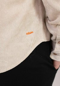 Beige kordfløyelskjorte med synlig ribbestruktur, med en oransje brodert detalj, knappede mansjetter og krum hemline. Svarte bukser i bakgrunnen.