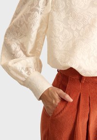 Blusa in pizzo crema con motivo floreale e maniche voluminose abbinata a pantaloni in velluto a coste color ruggine con tasche laterali e pieghe.