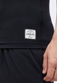 Κοντινή λήψη του χεριού ενός ατόμου κοντά σε μαύρο ριμπ πλεκτό πουκάμισο με λευκή ετικέτα Nike Athletic Club ραμμένη στο τελείωμα.