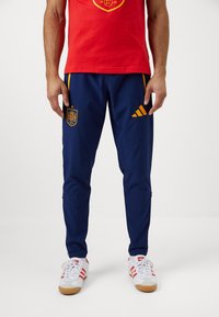 Muškarac nosi tamnoplave sportske hlače s narandžastim grbom španske nogometne reprezentacije i Adidasovim logotipom, u kombinaciji s bijelim i crvenim tenisicama.