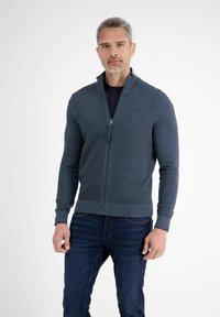 Mężczyzna z siwymi włosami, ubrany w ciemnoniebieski sweter na zamek błyskawiczny na czarnej koszulce oraz ciemnoniebieskie dżinsy, stojący na białym tle.