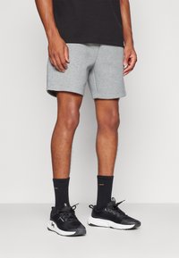 Uomo che indossa pantaloncini grigi, calzini neri e scarpe sportive nere con suola bianca, in piedi davanti a uno sfondo chiaro uniforme.