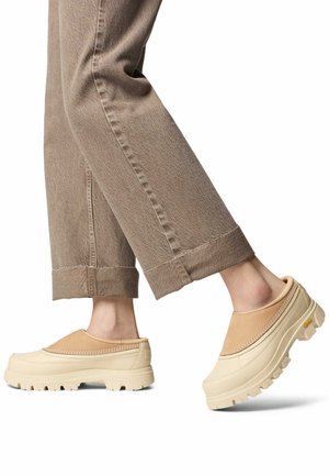 Fødder iført beige slip-on træsko med tykke såler og flettet kant, kombineret med opbukte brune bukser mod en hvid baggrund.