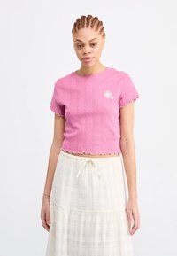 Rózsaszín, rövid ujjú crop top, texturált csíkokkal, hullámos szegéllyel és kis virágmintával a bal mellrészen. Fehér szoknyával viselve.
