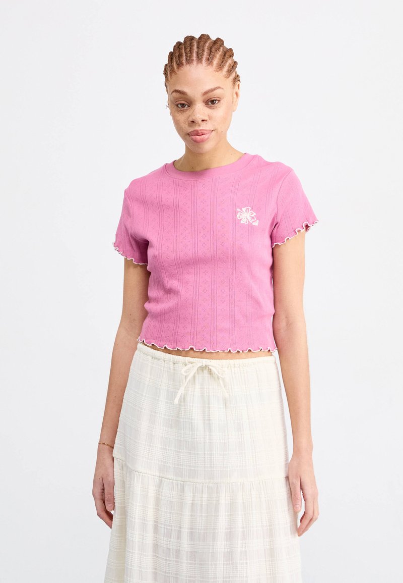 Rózsaszín, rövid ujjú crop top, texturált csíkokkal, hullámos szegéllyel és kis virágmintával a bal mellrészen. Fehér szoknyával viselve.