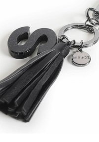 Llave negra con un acabado brillante en forma de la letra "S" y un fleco de cuero, con un anillo plateado y una pequeña etiqueta grabada con "MIRIADE."