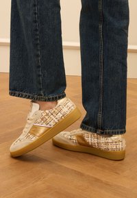 Beige Sneaker aus gewebtem Stoff-Obermaterial mit metallischen Goldakzenten. Gummisohle und Schnürdesign. Jeans mit fransigem Saum.