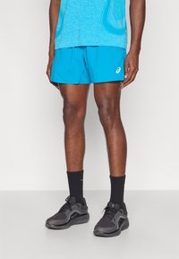 Τιρκουάζ αθλητικό σορτς με ελαστική μέση και λογότυπο. Μαύρα παπούτσια με υφασμάτινη λεπτομέρεια και μαύρες κάλτσες με πορτοκαλί λεπτομέρειες.