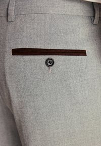 Pantalones en espiga grises con un bolsillo de acento de terciopelo marrón y un botón negro. La tela tiene una textura suave y un acabado ajustado.
