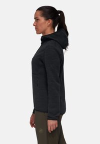 Dark gray hooded jacket with textured fabric, zippered chest pocket, and logo on the left sleeve. Paired with olive green pants.