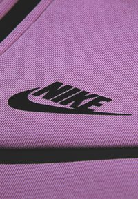 Μωβ ύφασμα με ραβδωτή υφή που παρουσιάζει ένα επιβλητικό μαύρο λογότυπο της Nike. Ο σχεδιασμός τονίζει το δυναμικό σχήμα του λογο τύπου σε αντίθεση με το φόντο.