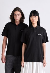 Dos modelos vistiendo camisetas polo negras de Lacoste, uno combinado con jeans oscuros y el otro con una falda blanca plisada, de pie frente a un fondo liso.