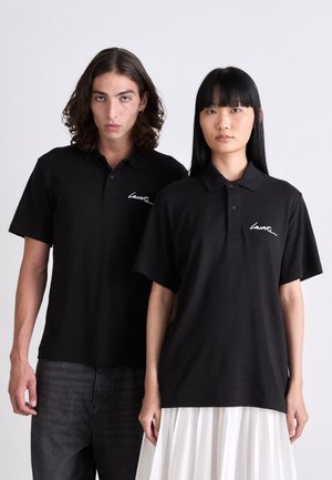 Două modele îmbrăcate în tricouri polo negre Lacoste, unul asortat cu blugi închisi la culoare și celălalt cu o fustă plisată albă, stând pe un fundal simplu.