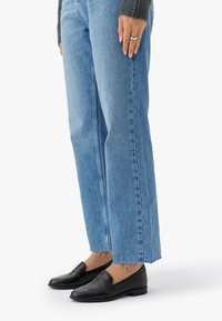 Des mocassins en cuir noir associés à un jean droit bleu clair. Les jeans ont des ourlets effilochés ; les mocassins présentent un faible talon et une finition lisse.