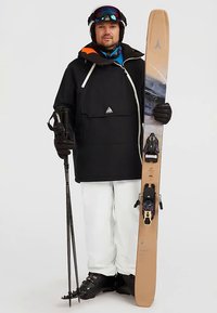 Uomo con giacca nera, pantaloni bianchi e attrezzatura da sci, che tiene sci beige e bastoni da sci su sfondo uniforme.