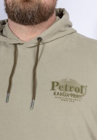 Felpa beige con cappuccio e cordino, dotata di un logo verde con la scritta "Petrod Kahua Trail". Il materiale sembra morbido con una finitura texture.