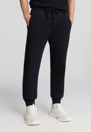 Pantaloni sportivi neri con vita elasticizzata e cordino, dotati di tasche laterali e polsini alle caviglie, abbinati a scarpe da ginnastica bianche.