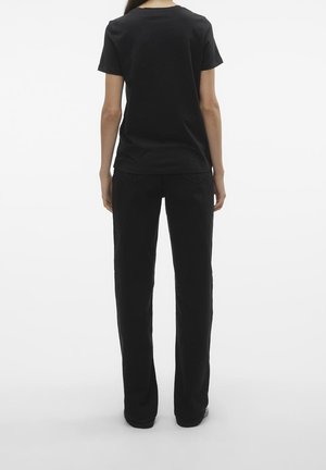 Persoon staande met de rug naar de camera, dragen een effen zwart T-shirt met korte mouwen en zwarte rechte broek tegen een witte achtergrond.