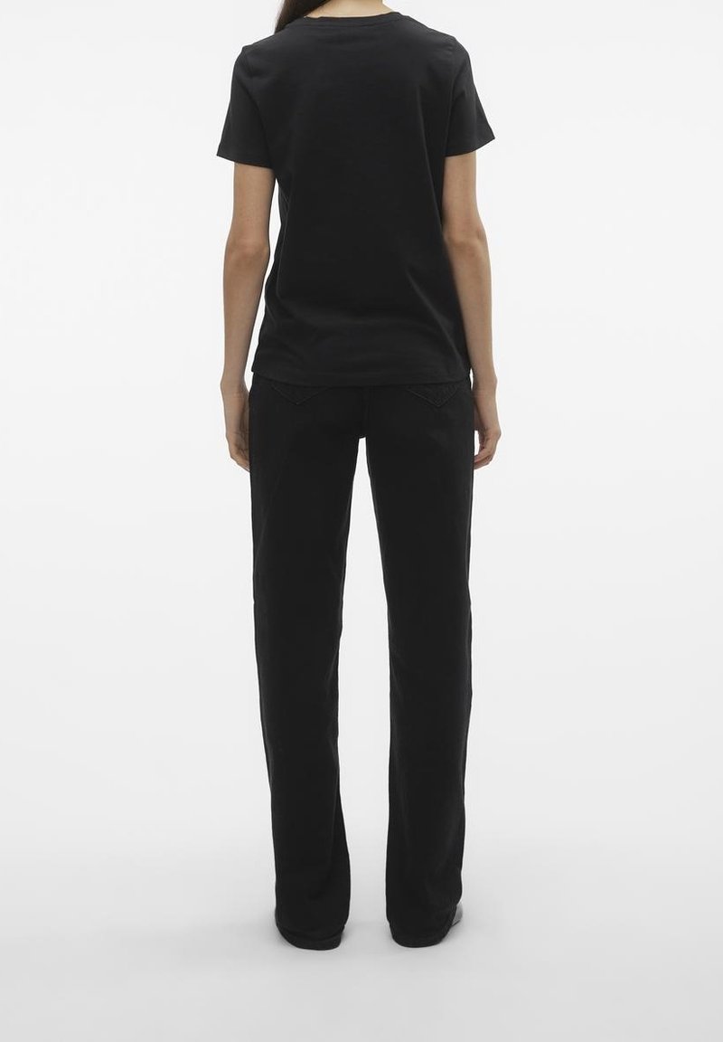 Personne debout de dos portant un t-shirt noir uni à manches courtes et un pantalon noir à jambes droites sur un fond blanc.