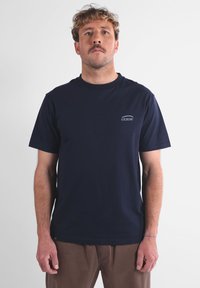 Homme avec une moustache portant un t-shirt Oxbow bleu marine et un pantalon marron, debout devant un fond blanc uni.