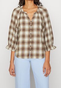 Brun och krämrandig skjorta med knappar fram, rund halsringning och puffärmar. Tillverkad av ett lätt tyg med en mjuk textur.