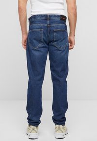 Jeans de color azul oscuro con un corte recto, diseño de cinco bolsillos y detalles en el bolsillo trasero. Combinados con zapatillas de tonos neutros.