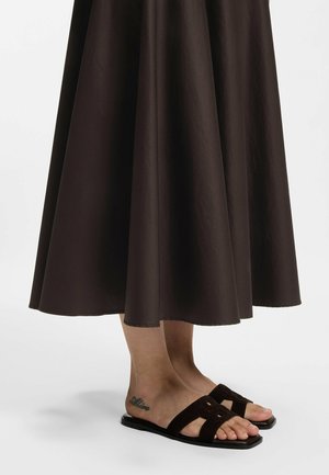 Feet wearing black flat sandals paired with a dark brown midi skirt, showing a tattoo on the ankle reading "Aline."