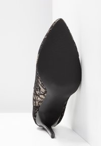 Chaussure à talon haut noire à bout pointu avec superposition de dentelle, vue du côté de la semelle reposant contre un mur blanc d'angle.