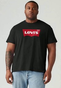 Tricou negru Levi's purtat de un bărbat cu antebrațul stâng tatuat și blugi de culoare albastru deschis, stând în fața unui fundal simplu de culoare deschisă.
