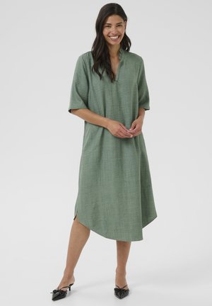 Green linen dress with short sleeves, a v-neck collar, and side slits, featuring a relaxed fit and a slightly tapered hem. Accented with smooth texture.