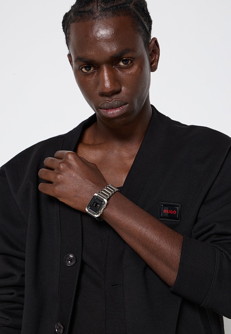 Cardigan negro con una tela texturizada, que presenta un parche del logo de Hugo en rojo y negro. Reloj plateado con una esfera cuadrada y banda de metal, llevado en la muñeca.