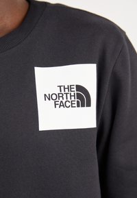 Sudadera negra con un parche cuadrado blanco que presenta el logo de "THE NORTH FACE" en texto negro y un gráfico curvado, fabricada en material de algodón suave.